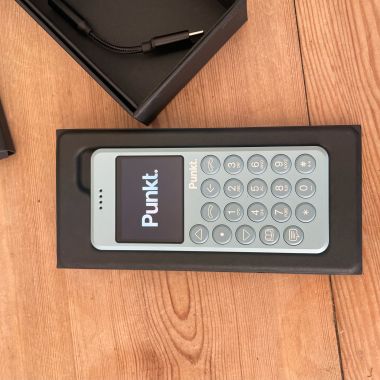 A Punkt MP02 light blue minimalist phone displayed in its packaging on a wooden surface, with a USB-C charging cable placed in a separate black box.