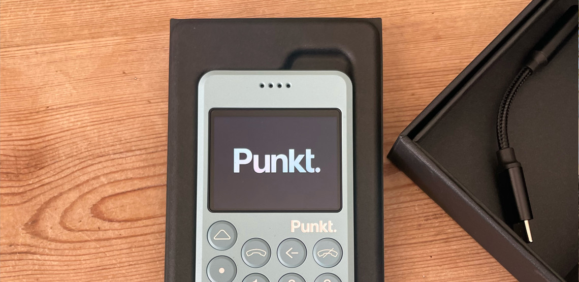 A Punkt MP02 light blue minimalist phone displayed in its packaging on a wooden surface, with a USB-C charging cable placed in a separate black box